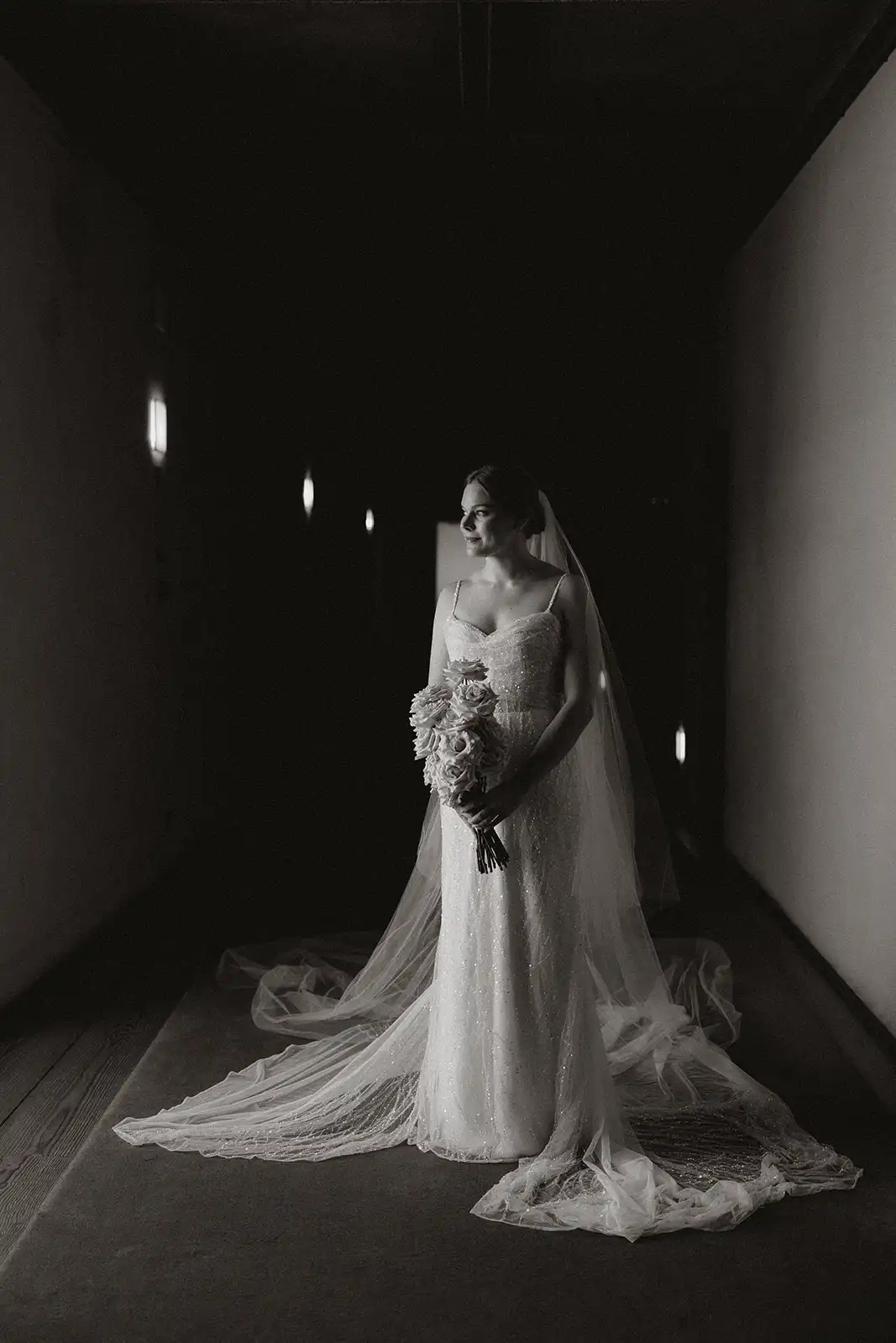 Bride illuminated by the light from the window in a black-and-white photo with a black-saturated background, in the corridors of the old monestary of Amares, Portugal.