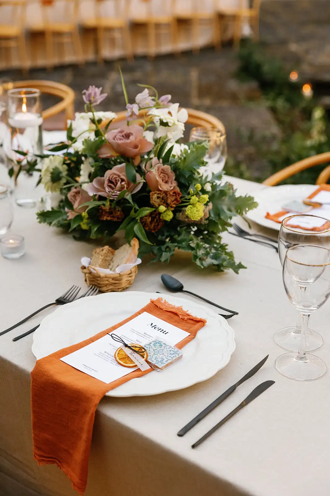 Close up of the wedding dinner table setup, neutral colors, orange napkin, black mate cutlery, litle basket to the bread. Flower arrangement centrepiece in dusty pink shades.