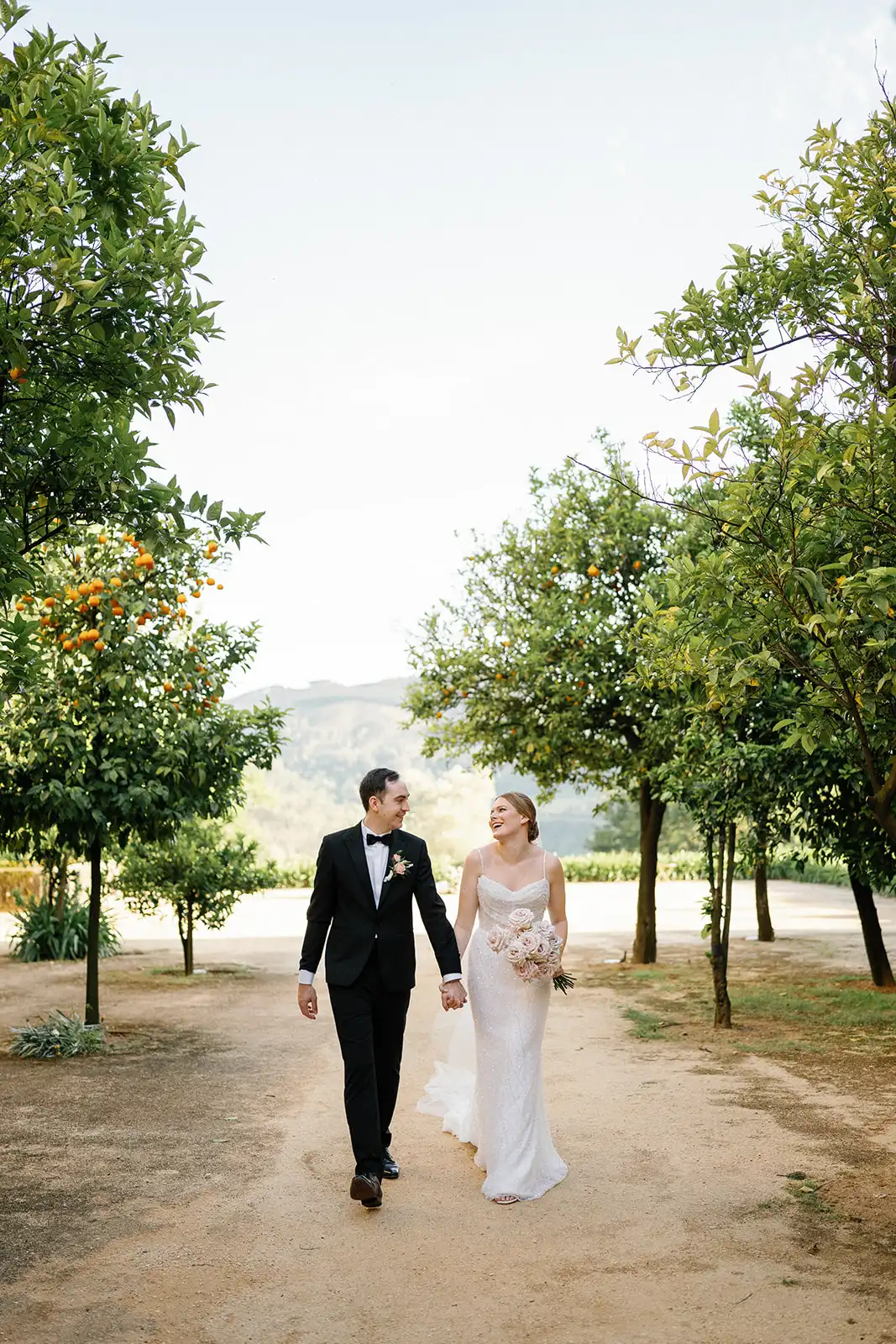 Bride and groom, in their wedding attire, walking hand in hand as they look at each other and smile, surrounded by the orange trees at the Pousada de Amares, with the Minho landscapes as background.