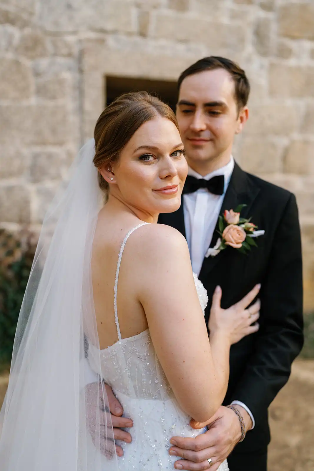 Bride and groom posing at the stone facade of Pousada de Amares. The bride looks directly into the camera, while the groom, who is slightly out of focus in the background, looks at the bride.