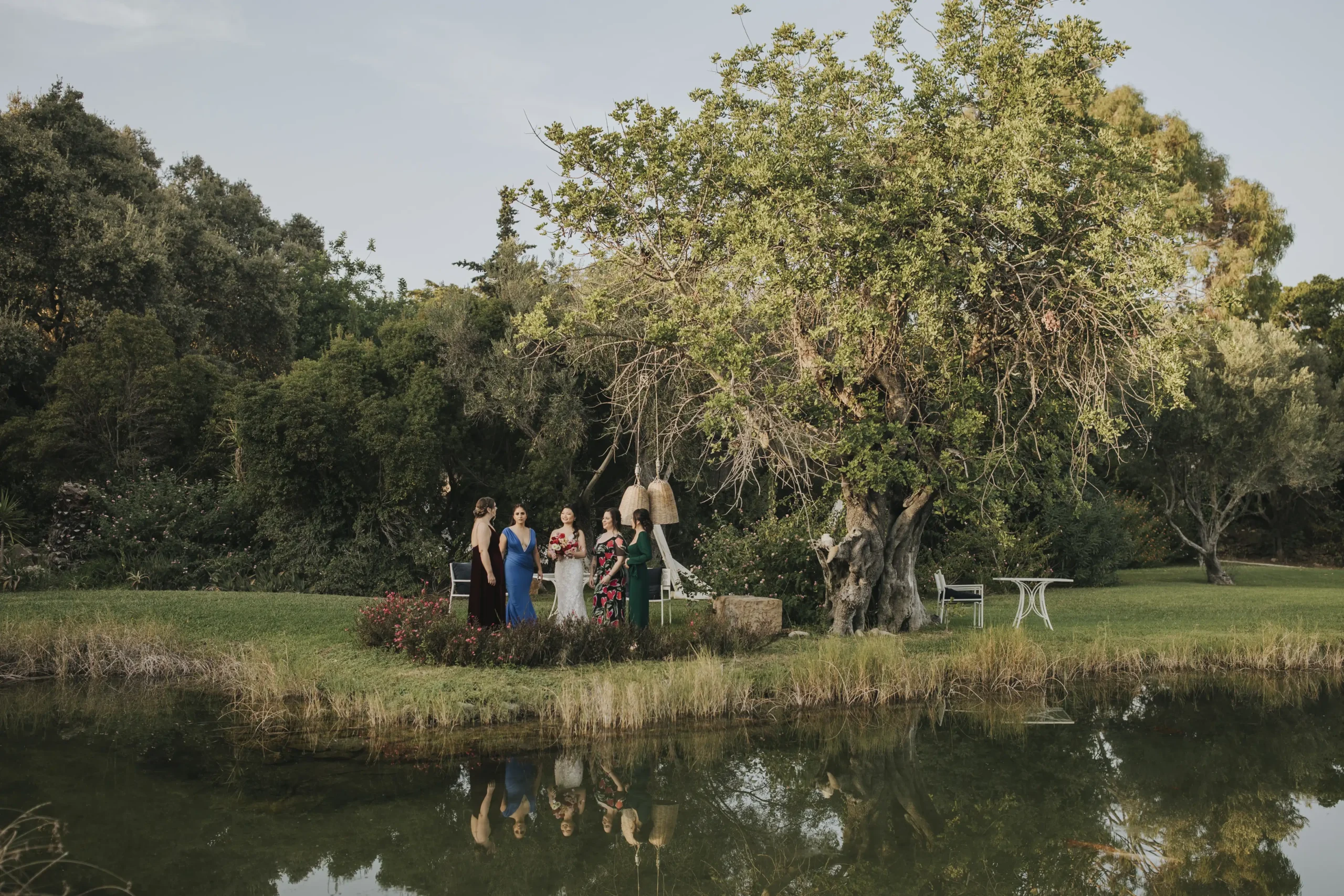Candid moment of the bride with bridesmaids in a beautiful corner of the venue, marked by the imposing tree and the lake that surrounds it, giving it the feel of an island. Portugal intimate wedding in Algarve.