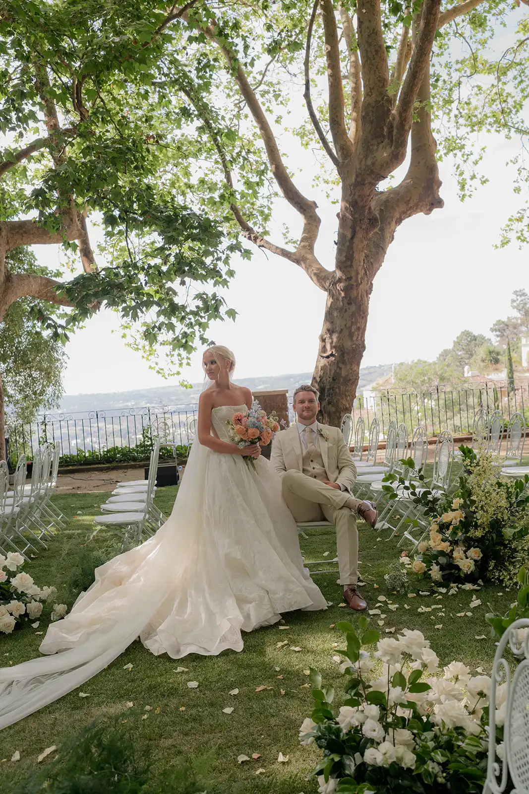 The bride and groom posing at ceremony location, surrounded by trees. A garden-style ceremony with iron chairs and grass. A color palette of pink, peach, and yellow. View of Sintra.