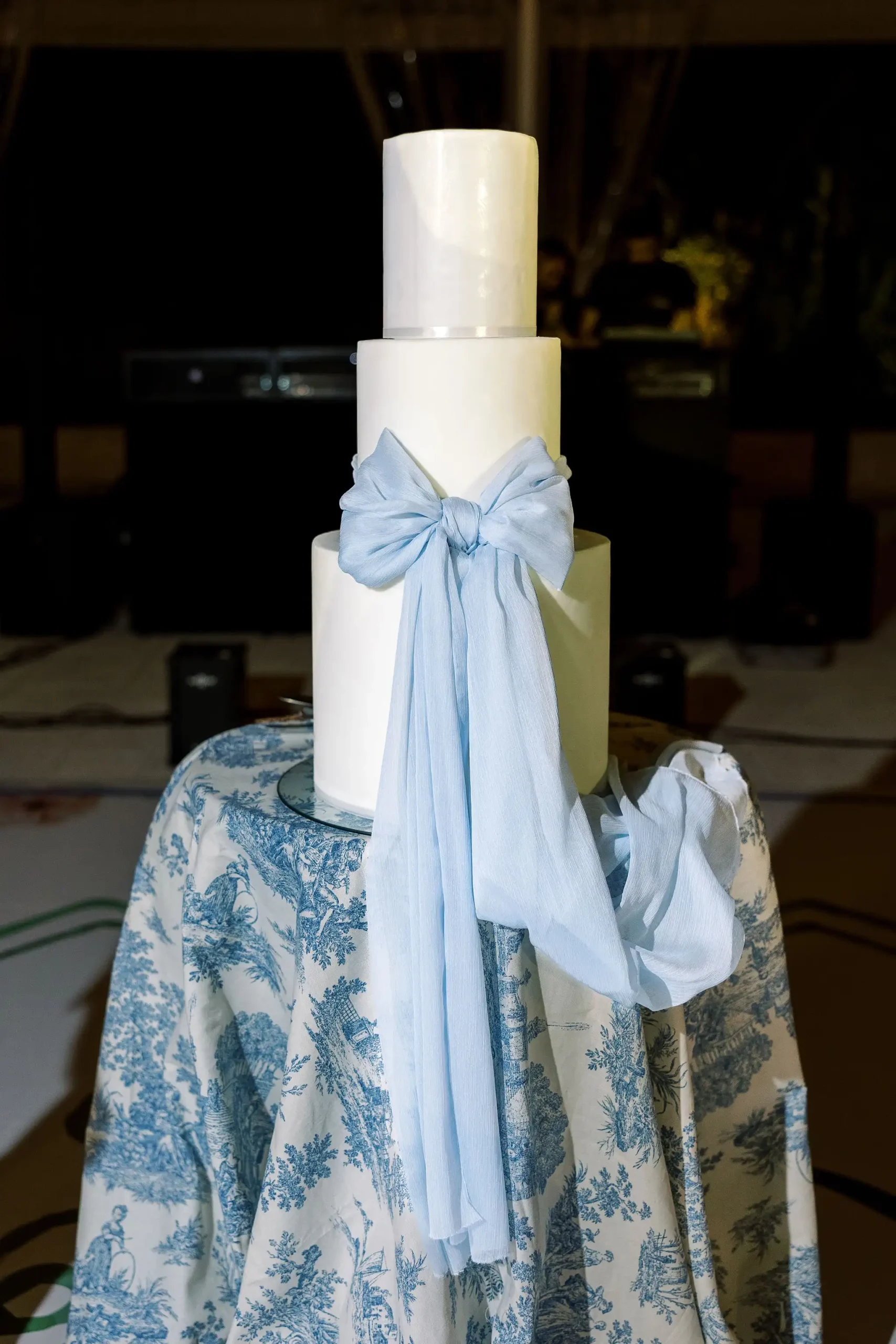 Cake table with blue Toile-de-Jouy tablecloth and white three-tier wedding cake with blue fabric bow as decor.
