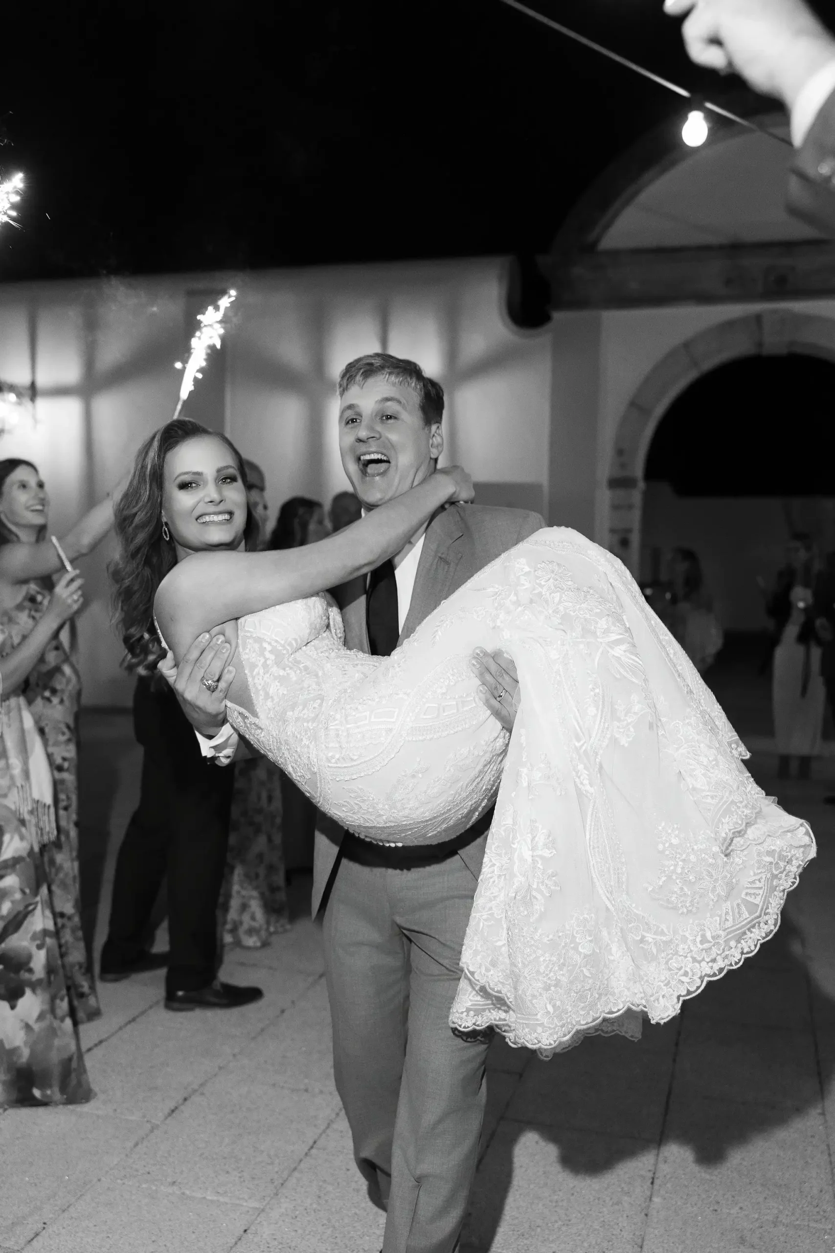 Groom carrying his bride in his arms as they walk down the aisle lined with guests holding lit sparklers. The bride and groom leave the event.