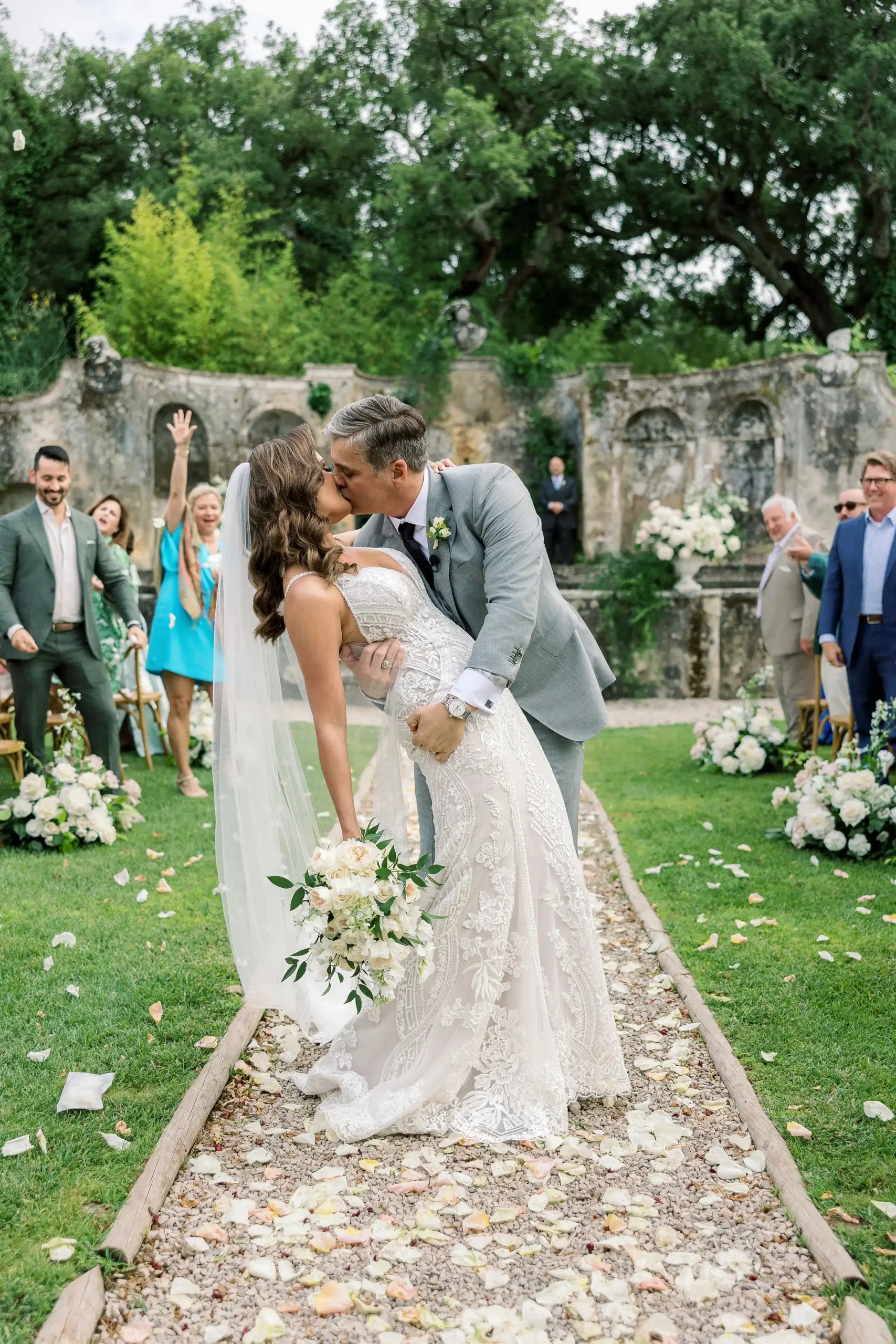 Bride and groom kiss in the aisle of the ceremony full of petals. Guest celebrating. Wedding ceremoni in historic garden.