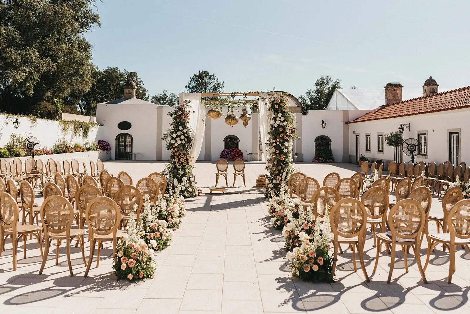 Indian wedding ceremony decor in a destination wedding in Portugal. Mandap with fabrics, gold lamps and neutral color flowers in a historic country retreat patio. Aisle with neutral flower arrangements and wooden classic chairs.