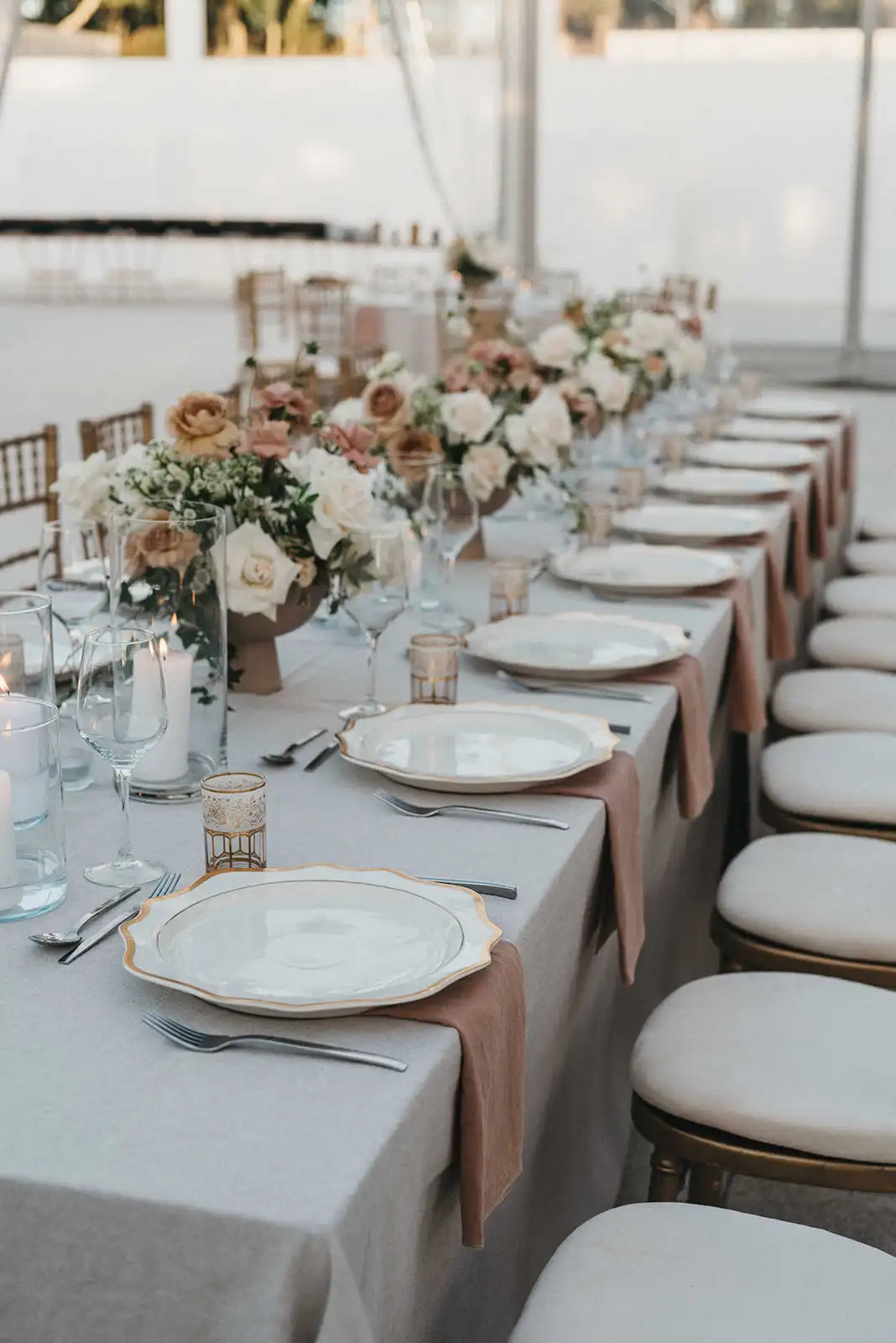Wedding dinner long table decor with neutral tones. Off-white, gold and dusty pink tablescape with gold details. Pilar candle centerpieces and beige and pink flower arrangements.
